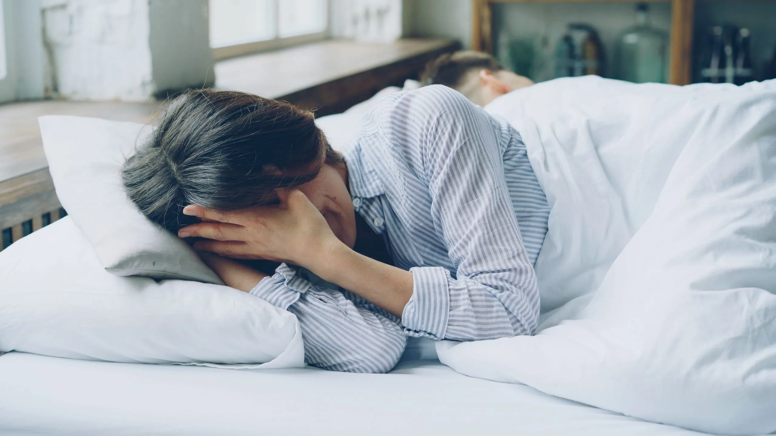 Erschöpfte Frau liegt nachdenklich im Bett, während ihr Partner abgewandt schläft – Symbolbild für Beziehungsprobleme nach der Geburt.