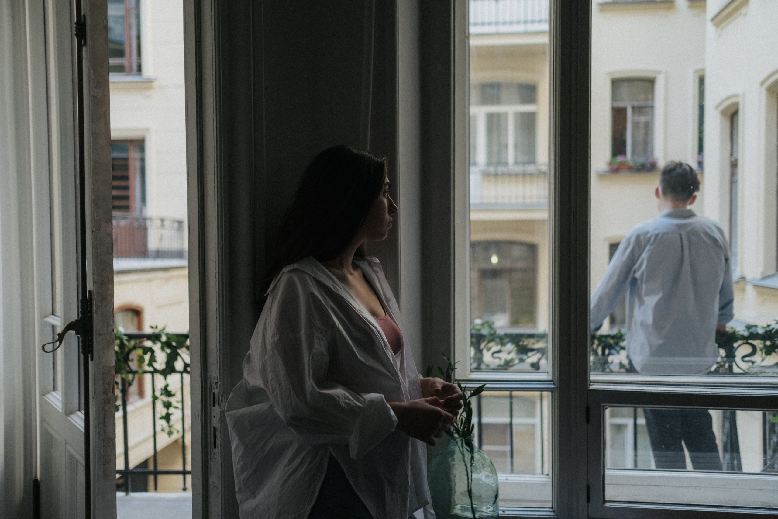 Streit nach der Geburt: Eine Frau steht nachdenklich im Zimmer, während ihr Partner sich auf dem Balkon abwendet.
