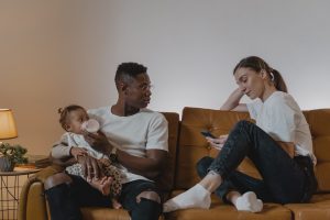 Streit nach der Geburt: Ein Vater füttert das Baby auf dem Sofa, während die Mutter erschöpft und zurückgezogen auf ihr Smartphone schaut.